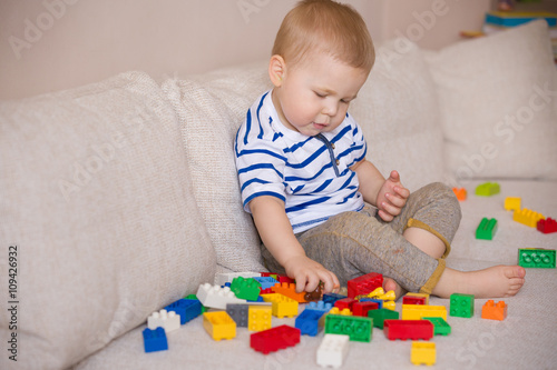 Cute little toddler boy in a striped shirt playing with colorful plastic blocks on the sofa indoors. child having fun and building out of bright constructor bricks. Early learning. Creative.
