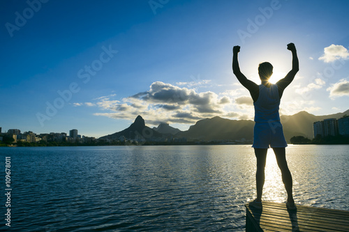 Silhouette of athlete standing with arms raised at sunset in front of Rodrigo de Freitas lagoon in Rio de Janeiro, Brazil