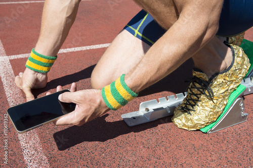 Athlete crouching at the starting line of a running track listening to motivational music on headphones from his mobile phone armband with Brazil colors wristbands 