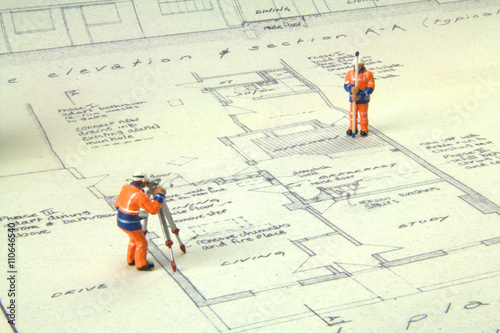 Two miniature figures in high-vis uniform with a theodolite survey a house blueprint