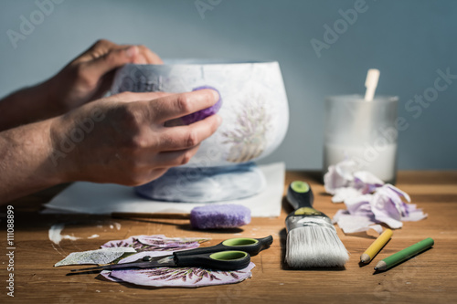 Decoupage hobbyist hands decorating a vase with lavender pattern - some artistic supplies on a table. 
