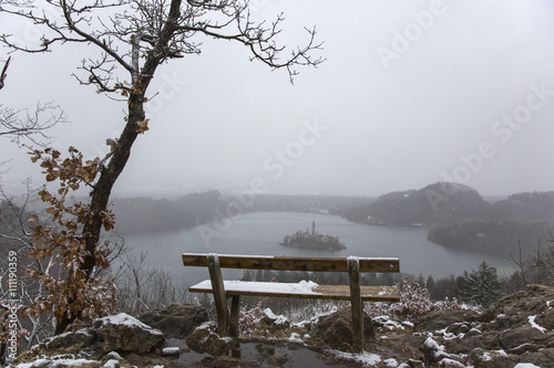 Snowy bench with a view on island in the middle of Lake Bled. Slovenia.