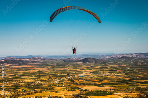 A paraglider flies over picturesque fields in the background of blue sky