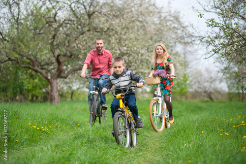 Happy boy on a bicycle in front of and behind its parent in the spring blooming garden riding bikes. Family having fun against the fresh greenery