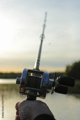Male hand holding a round fishing reel attached to a fishing rod with blurred sea nature on the background at sunset time. 