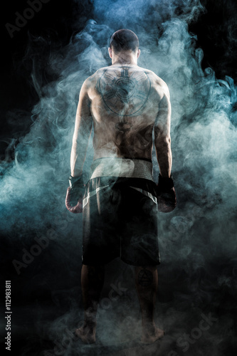 Muscular kickbox or muay thai fighter with tattoo standing back in smoke.