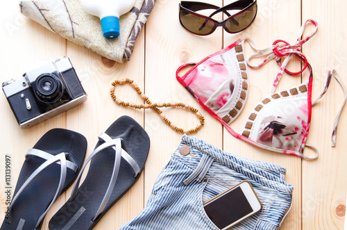 Summer accessories for travel or vacation concept. Vintage camera, denim shorts glasses, bikini, flip flop, beach towel, sunburn lotion, mobile on wooden background.