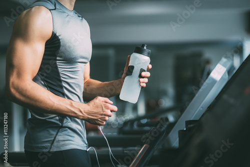Unrecognizable young man in sportswear running on treadmill at gym and holding bottle of water