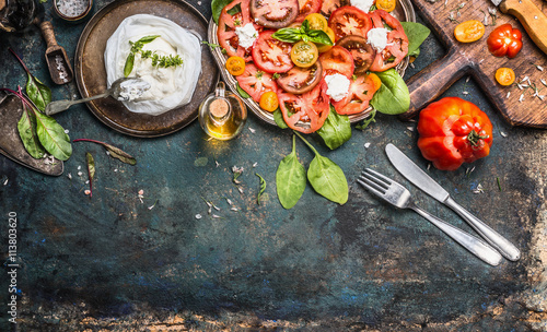 Tomatoes and mozzarella salad, preparation on dark aged rustic background, top view. Italian lunch with tomatoes and mozzarella, cutlery and cooking ingredients. Italian food concept