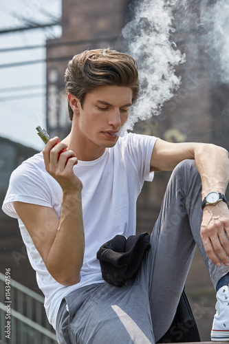 Junger Mann in T-Shirt und Jeans sitzt e-Zigarette rauchend an einer Bahnstrecke und bläst lässig eine weiße Dampfwolke.
