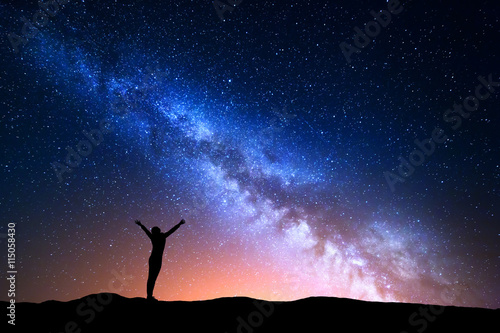 Landscape with blue Milky Way and yellow light. Colorful night sky with stars and silhouette of a standing sporty girl with raised-up arms on the hill on the background of beautiful universe.