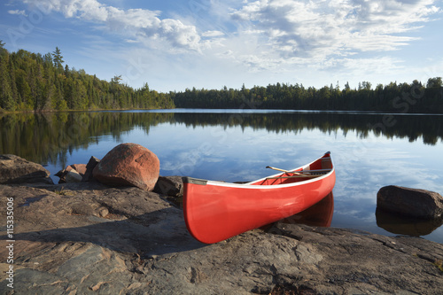 Red canoe on rocky shore of calm lake with pine trees