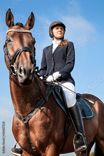 Portrait of serious beautiful young girl jockey in uniform sitting on a horse against blue sky and yellow field and looking forward on a sunny day. Equestrian sport - dressage.