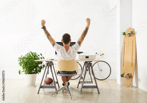 Tattooed lucky freelancer in front of his working space, surrounded with his hobby toys longboard, vintage bicycle and green plant, stretching his hand in air while making break