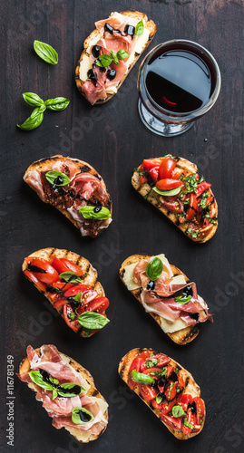 Brushetta set with glass of red wine. Variety of small sandwiches with prosciutto, tomatoes, cheese, herbs and balsamic creme on dark wooden background, top view