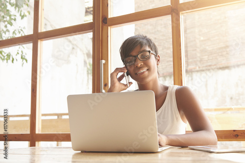 Beautiful young freelancer talking on phone with her clients. Dark-skinned girl in glasses with brunette short hair looks happy and content to do her job ideally and working with laptop in a caf?.