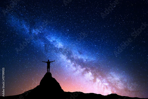 Night landscape with Milky Way. Silhouette of a standing man with raised up arms on the top of mountain. High rocks, mountain peak. Beautiful Galaxy. Universe. Blue night starry sky and city lights