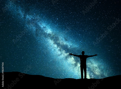 Night colorful landscape with green Milky Way and silhouette of a happy man with raised up arms on the hill. Beautiful Universe. Travel background with starry sky. 