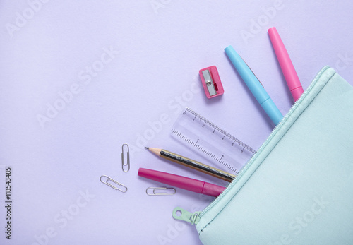 An aerial view of a blue pencil case with stationery spilling out on to a pastel purple background, forming a page border