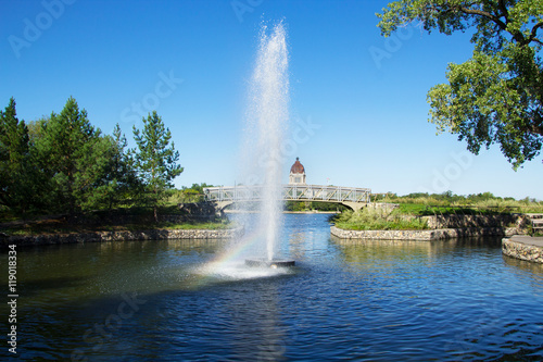Early morning photo of a fountain in Regina Saskatchewan's Wascana Park with a rainbow in the spray. Landscape view with a footbridge and the provincial legislature in the distance.