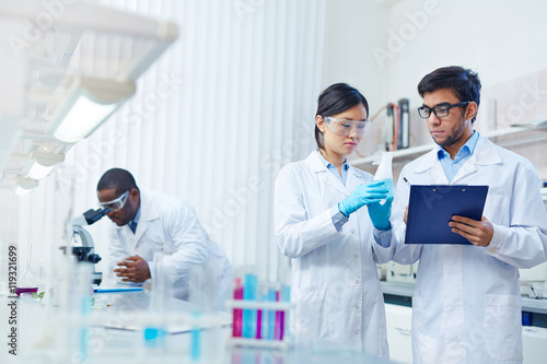 Concentrated Asian female laboratory scientist in safety goggles holding flask with blue liquid showing it to Latin-American colleague writing down results. African-American scientist in background.