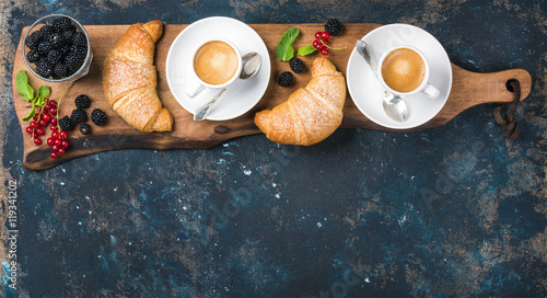 Breakfast set. Freshly baked croissants with garden berries and coffee cups served on rustic wooden board over dark grunge plywood backdrop, top view, copy space