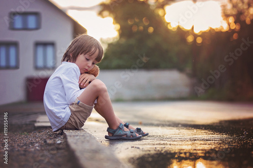 Sad little boy, sitting on the street in the rain, hugging his t