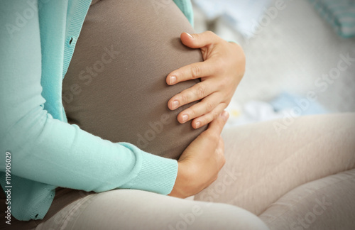 Pregnant woman holding hands on belly and sitting on sofa in room