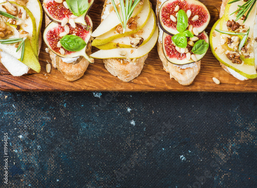 Crostini with pear, ricotta cheese, figs, nuts and fresh herbs. Breakfast toasts or snack sandwiches on rustic wooden board over dark blue grunge plywood background. Top view, copy space