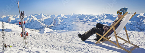 Girl sunbathing in a deckchair near a snowy ski slope in winter, pair of skis in the snow, panorama of alpine mountains with copy space in The Alps, France