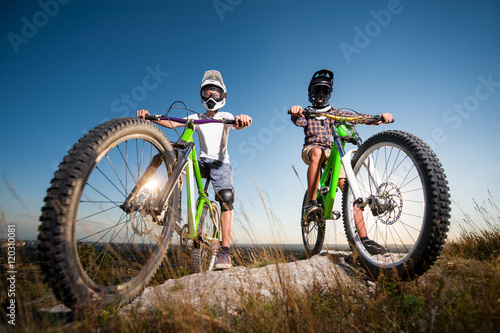 Two cyclists wearing in helmets and glasses standing with the mountain bikes on the top of the mountain under blue sky. Men looking into the camera. Bottom view