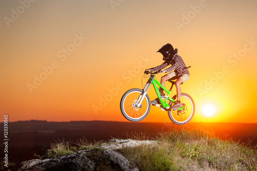 Guy high jump on a mountain bike on the top of hill against evening sky with bright sun. Cyclist is wearing sportswear helmet and glasses. Extreme freeride. Sunset