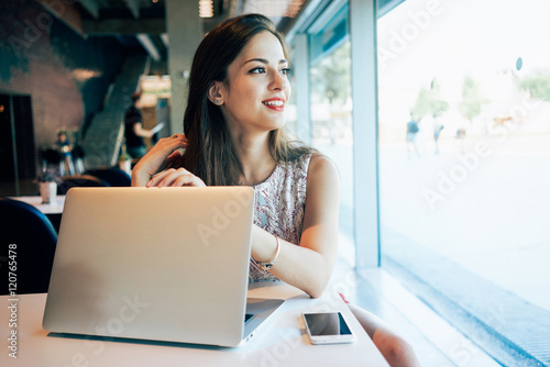 A caucasian female freelancer is looking aside while sitting in a cafe with a portable computer and a smartphone on a table. Smiley european girl sitting with electronic devices in a co-working.