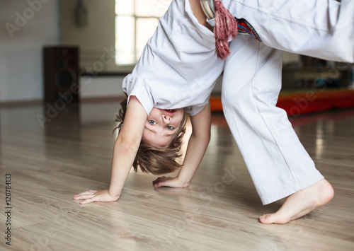 Cute boy in white clothes practicing capoeira (brazilian martial art that combines elements of dance, acrobatics and music) in gym 