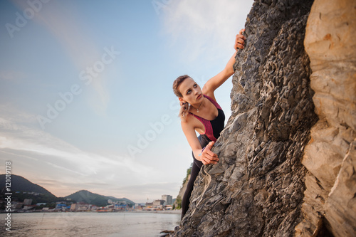 Portrait of young female rock climber overhanging cliff. Beautiful blue sky, city mountains on the background.