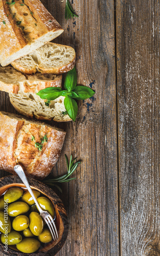 Green mediterranean olives in bowl and slices of freshly baked ciabatta over rustic wooden background. Top view, copy space, vertical composition
