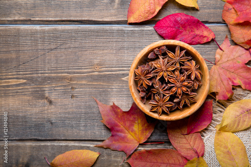 Star anise in a wooden bowl on a wooden table with colorful autumn leaves around. Top view with copy space.