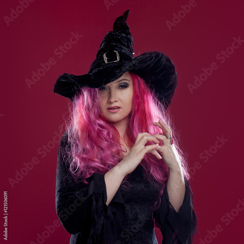 Attractive woman in witches hat and costume with red hair performs magic on pink background. Halloween, horror theme.