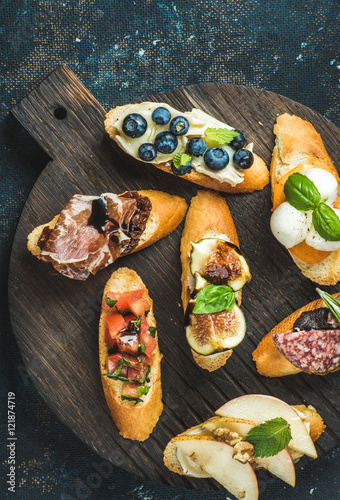 Italian crostini with various toppings on round wooden serving board over black plywood background, top view