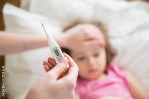 Close-up thermometer. Mother measuring temperature of her ill kid. Sick child with high fever laying in bed and mother holding thermometer. Hand on forehead. 