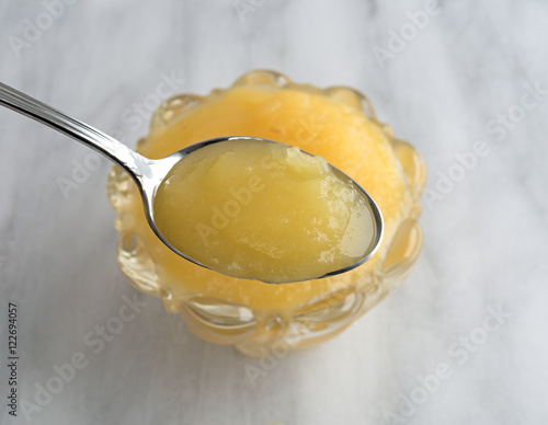 Spoon full of applesauce with a glass bowl underneath atop a marble table.