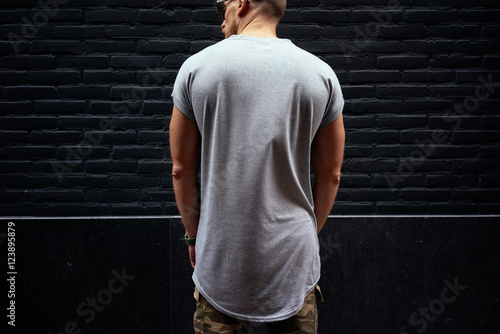 A cropped photo of a young man in a blank gray t-shirt standing with his back to the camera on a black brick wall background. Empty place for the logo or design. Mock up.