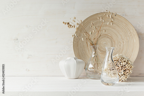 Soft home decor of glass vase with spikelets and wooden plate on white wood background. Interior.
