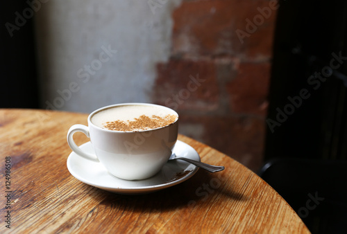 Cup of tasty latte is standing on the wooden textured table with the brick wall background. Latte is in the white big cup.