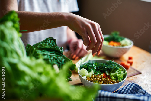 Female hand pours green onions in a bowl with green peas, cucumbers, carrots, lettuce and dill standing on a table
