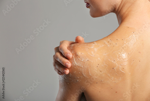 Young woman applying scrub on shoulder on grey background