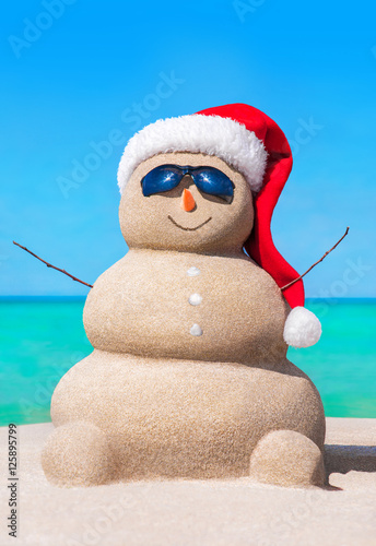 Sandy snowman in Christmas hat and sunglasses at tropical beach