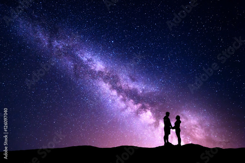 Milky Way with silhouette of people. Landscape with night sky with stars and standing man and woman holding hands on the mountain. Hugging couple against purple milky way. Beautiful galaxy. Universe 