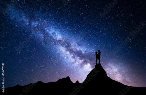 Milky Way with silhouette of people. Night landscape with starry sky. Standing man and woman on the top of mountain. Hugging couple against milky way. Beautiful galaxy. Universe. Travel. Bright stars