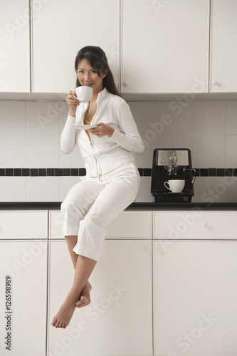 Woman sitting on counter in kitchen next to cappuccino machine, drinking coffee and looking at camera.